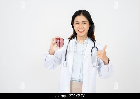 Attraktive Ärztin mit einem weißen, lächelnden Mantel und einem Apfel in der Hand. Fröhliche medizinische Ernährungsberaterin empfiehlt eine gesunde Ernährung für einen Patienten. Stockfoto