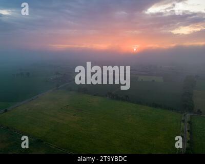 Dieses Bild fängt die ruhige Schönheit der Landschaft in der Abenddämmerung ein. Die untergehende Sonne blickt durch den trüben Himmel und strahlt ein warmes Leuchten aus, das im Kontrast zu den kühlen Schatten steht, die das Land umhüllen. Eine Nebeldecke verdeckt teilweise die Felder und fügt der Landschaft eine Schicht von Geheimnissen und Ruhe hinzu. Die fernen Häuser und Bäume werden zu Silhouetten gegen das schwindende Licht und markieren das friedliche Ende des Tages Die Dämmerung fällt auf die Landschaft. Hochwertige Fotos Stockfoto