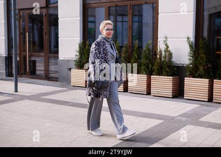 Stilvolle blonde, reife Frau mit kurzen Haaren trägt modisches mehrlagiges Winteroutfit, Mantel mit Tierdruck, grauer Wollanzug mit Sneakern, Spaziergänge in der Stadt Stockfoto