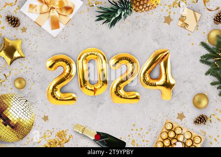 Neujahr 2024, kreative Idee. Goldene Ballons 2024 liegen auf einem grauen Betontisch mit Kaminen, Süßigkeiten, einer goldenen Discokugel, Champagner und einem Weihnachtsfest Stockfoto
