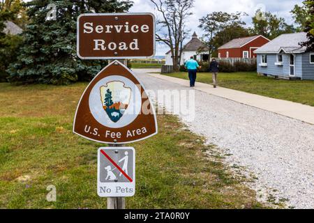 Ein Schild mit der Aufschrift „Service Road“, das das Pfeilspitzen-Logo des National Park Service und eine „U.S. Fee Area“-Beschriftung trägt, steht neben dem Weg zur Sleeping Bear Point Coast Guard Life-Saving Station Maritime Museum in der Nähe von Glen Arbor Township, Michigan, Vereinigte Staaten. Besucher gehen den Kiesweg zwischen den historischen Gebäuden der ehemaligen Rettungsstation entlang. Stockfoto