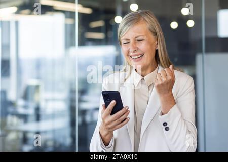 Glückliche Geschäftsfrau, die im Büro steht und auf das Handy blickt. Ich freue mich, die Nachricht und die gute Nachricht zu erhalten. Macht mit seiner Hand eine Siegesgeste. Stockfoto