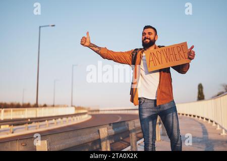 Der Mann trampt am Straßenrand und versucht, das Auto anzuhalten. Er hält Pappe mit Inschrift in der Hand. Stockfoto