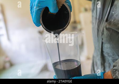 Nahaufnahme der Hände, die schwarze Farbe mit blauen Handschuhen in einen klaren Messbecher gießen Stockfoto