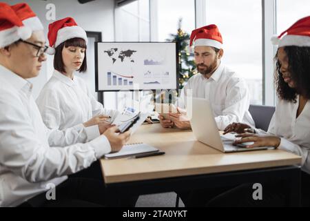 Multiethnische Geschäftskollegen treffen sich im Büro mit Plasmabildschirm und Weihnachtsschmuck. Gutaussehender bärtiger Teamchef Stockfoto