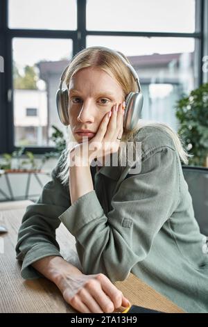 Vertikale Aufnahme einer jungen, nicht binären Person mit Kopfhörern, die Hand unter dem Kinn in die Kamera schaut Stockfoto