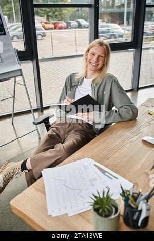 Fröhliche, nicht binäre Person, die am Schreibtisch sitzt und das Notebook in den Händen hält und in die Kamera lächelt, geschäftlich Stockfoto