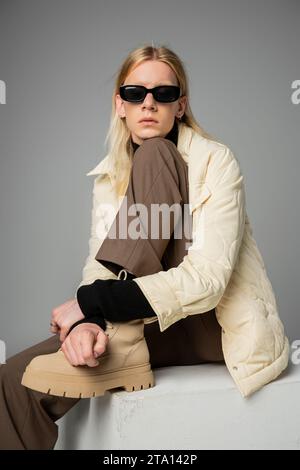 Gut aussehender androgyner Mensch in trendiger Winterkleidung, sitzt auf einem riesigen weißen Würfel, Modekonzept Stockfoto