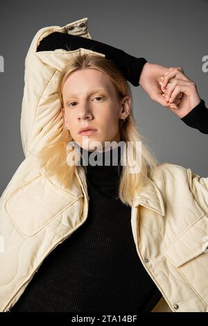 Attraktive androgyne Person im Winter stilvolle Jacke posiert auf grauem Hintergrund mit Armen über dem Kopf Stockfoto
