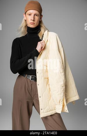 Gut aussehendes, nicht binäres Modell posiert mit stilvoller Winterjacke auf der Schulter und blickt in die Kamera Stockfoto