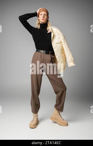 Gut aussehender androgyner Mensch in trendigem Winteroutfit mit Hand auf Kopf, Blick in die Kamera, Mode Stockfoto