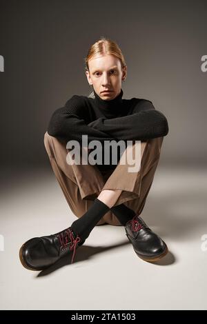 Vertikale Aufnahme einer jungen nicht binären Person mit schwarzem Rollkragen, die auf dem Boden sitzt und in die Kamera blickt Stockfoto