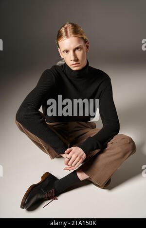 Ansprechende, nicht binäre Person mit rotem Haar und Pferdeschwanz, die mit überkreuzten Beinen auf dem Boden sitzt, Mode Stockfoto