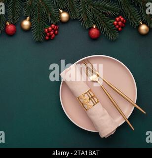 Weihnachtstisch. Leichte Keramikplatte, goldene Gabel, Messer. Tannenzweig und glänzende Kugeln auf grünem Hintergrund, Einladungsdesign. Neujahrs d Stockfoto