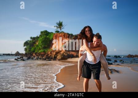 Ein Paar genießt einen Huckepack-Ritt bei Sonnenuntergang am tropischen Strand. Der Mann trägt eine freudige Frau auf dem Rücken, die entlang der Küste schlendert. Romantische Reisende am Meer Stockfoto