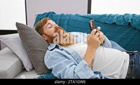 Gutaussehender bärtiger irischer rothaariger Mann, völlig entspannt und ernst, konzentriert auf die Benutzung seines Telefons, verloren in einer Welt der Internet-Technik, als er auf dem Sofa liegt Stockfoto