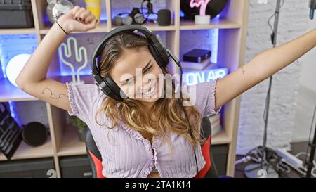 Junge hispanische Frau, eine attraktive Streamer mit gewinnendem Lächeln, die fröhlich zu Musik tanzt, während sie im dunklen Spielzimmer Spiele streamt Stockfoto
