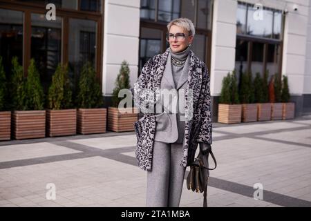 Stilvolle blonde, reife Frau mit kurzen Haaren trägt modisches mehrlagiges Winteroutfit, Mantel mit Tiermuster, grauen Wollanzug mit Rollkragen und Brille Stockfoto