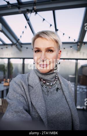 Selfie-Aufnahme einer schönen Reifen blonden Frau mit kurzen Haaren, die elegante graue Kleidung mit Accessoires trägt. Nahaufnahme Porträt von elegantem Mittelteil Stockfoto