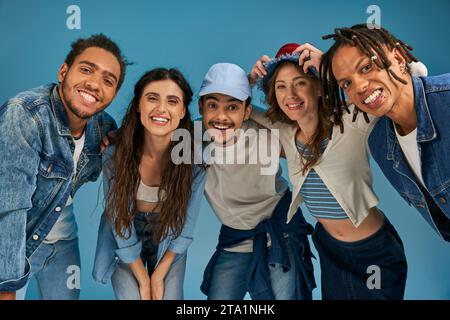 Fröhliche, multiethnische Freunde in trendiger Streetwear lächeln in der Kamera vor blauem Hintergrund, urbaner Stil Stockfoto