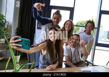 Fröhliche Frau mit Smartphone, die Selfie mit multiethnischen Team-Gesten im modernen Büro macht Stockfoto