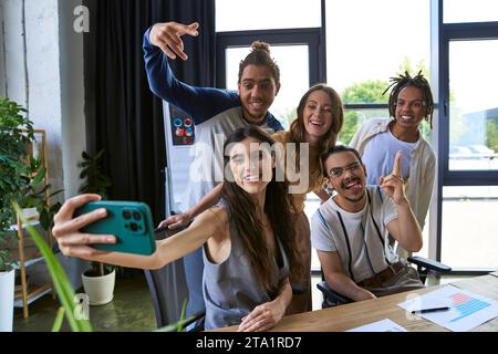 Lächelnde Frau, die Selfie auf dem Smartphone mit glücklichen multikulturellen Kollegen im modernen Büro macht Stockfoto