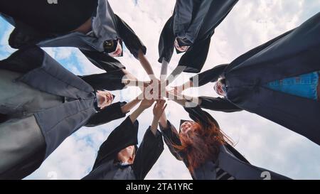 Gruppe glücklicher erfolgreicher Absolventen in akademischen Hüten und Gewändern, die im Kreis stehen und ihre Hände zusammenlegen. Team der Hochschule oder Universität Stockfoto