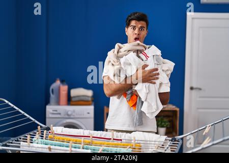 Hispanischer Mann, der Kleidung an der Wäscheleine im Schockgesicht hängt, skeptisch und sarkastisch aussah, überrascht mit offenem Mund Stockfoto