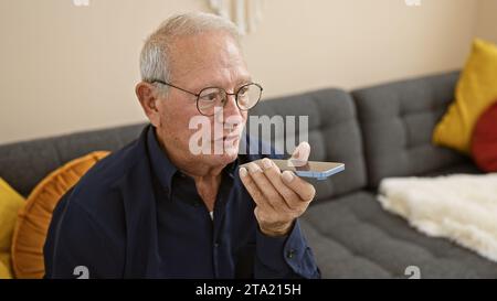 Ein ernstzunehmender älterer Mann mit weißem Haar, vertieft in die Technologie eines Smartphones, sendet von seinem Wohnzimmer-Sofa zu Hause eine Audiobotschaft Stockfoto
