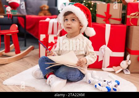 Ein entzückender hispanischer Junge, der den weihnachtsmann-Brief schreibt, sitzt zu Hause am weihnachtsbaum auf dem Boden Stockfoto