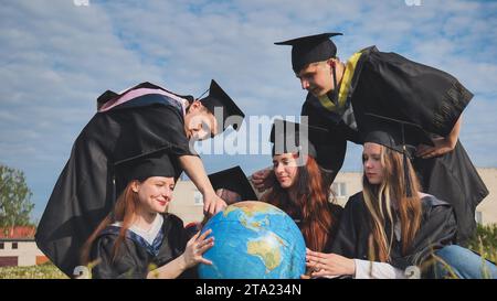 Absolventen in schwarzen Gewändern untersuchen einen geografischen Globus, der auf dem Gras sitzt. Stockfoto