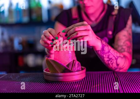 Nahaufnahme einer alternativen Barkeeperin, die einen Margarita-Cocktail mit Teufelshörnern in einer Nachtbar mit Neonlichtern dekoriert Stockfoto