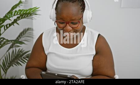 Lächelnde afroamerikanerin sitzt im Wartezimmer, hört über Kopfhörer gefühlvolle Lieder auf dem Touchpad, Chefin schaukelte Zöpfe und Brillen Stockfoto