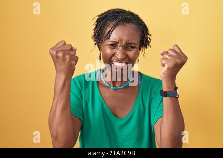 Afrikanische Frau mit Dreadlocks, die über gelbem Hintergrund stehen, wütend und wütend, die Fäuste hochziehen, frustriert und wütend, während sie vor Wut schreien. Wut und A Stockfoto