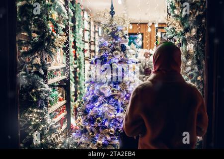 Rückansicht einer Frau, die nachts auf der Straße der Stadt eine zu Weihnachten dekorierte Ladenpräsentation ansieht. Weihnachtszeit Stockfoto