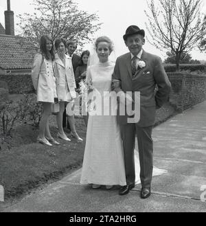 1960er Jahre, historisch, Hochzeit, ein stolzer Vater, der in einem Anzug mit Nelkenblume und Hut elegant aussieht, begleitet seine Tochter, die Braut, mit einem glücklichen Hufeisen, eine Kirche hinauf, einen Pfad, beobachtet von zwei jungen Frauen in Miniröcken, England, Großbritannien. Stockfoto