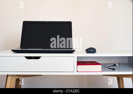 Leerer, offener Laptop mit Mockup und schwarzem Bildschirm auf weißem Schreibtisch. Maus. Buch und Kabel. Moderner Arbeitsbereich Stockfoto