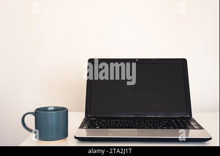 Leerer, offener Laptop mit Mockup und schwarzem Bildschirm auf weißem Schreibtisch. Tasse mit Kaffee. Minimalistischer Arbeitsbereich Stockfoto