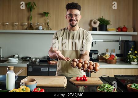 Mittelgroße Aufnahme eines lächelnden jungen männlichen Influencers aus dem Nahen Osten, der in die Kamera blickt, die einen Karton mit Eiern hält, während er in der Küche posiert Stockfoto