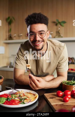 Junger Influencer aus dem Nahen Osten, der in die Kamera blickt und eine Gabel mit einem Stück Omelett auf dem Küchentisch mit Geschirr und Gemüse hält Stockfoto