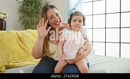 Eine reizende Mutter hält selbstbewusst die kleine Tochter in den Armen und genießt eine fröhliche, zwanglose Unterhaltung auf dem Sofa. Herzerwärmender Familienmoment voller fröhlichem Lächeln Stockfoto
