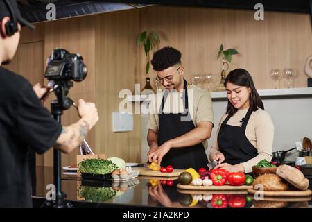 Mittelgroße Aufnahme eines jungen Mannes aus dem Nahen Osten und eines hispanischen Mädchens, der an der Küchentheke steht und Gemüse schneidet, während er von einem Kameramann gefilmt wird Stockfoto