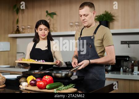 Mittelgroßer Schuss einer jungen hispanischen Frau und eines kaukasischen Küchenchefs, der am Küchenherd steht und gehacktes Gemüse in der Pfanne frittiert Stockfoto