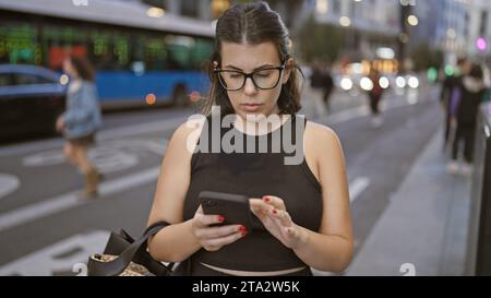Schöne brünette hispanische Frau in Brille, vertieft in ernsthafte Smartphone-Gespräche auf madrids städtischen Straßen Stockfoto