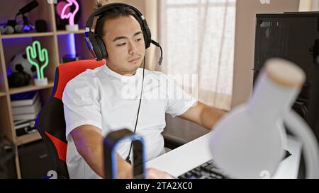 Konzentrierter junger chinese, ein Streamer für Technik, der sich ernsthaft in ein futuristisches digitales Spiel in der Nacht im Spielzimmer vertieft hat. Stockfoto