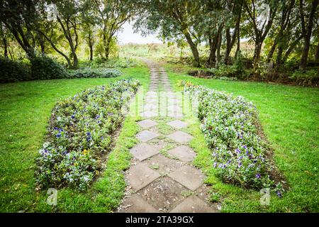 Ein malerischer Steinweg, gesäumt von einer Vielzahl von bunten Blumen, schlängelt sich durch einen üppigen, grünen Park, der von hoch aufragenden Bäumen gesäumt ist Stockfoto