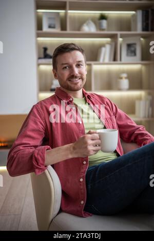 Positiver glücklicher Mann, der lächelnd eine Tasse Tee hält, der im Stuhl sitzt und die Kamera im gemütlichen Zuhause sieht. Stockfoto