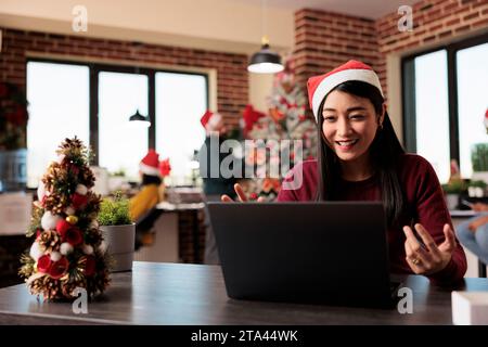 Lächelnder Angestellter mit weihnachtsmütze, der das Projekt bei Telefonkonferenzen mit Laptop in einem Weihnachtszimmer leitet. Fröhliche Arbeitnehmerin mit Online-Kommunikation am Arbeitsplatz während der Neujahrsferien Stockfoto