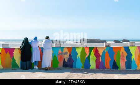 Essaouira, Marokko - 17. September 2022: Marokkanische Familie in traditioneller Kleidung, die sich an einer bunten Mauer stützt, um den Atlantischen Ozean an der Küste zu beobachten Stockfoto