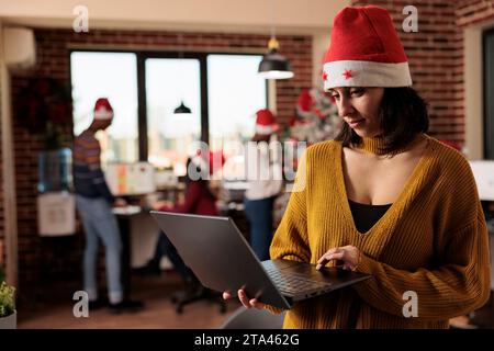 Weiße Angestellte mit weihnachtsmann-Hut und Laptop im weihnachtlich dekorierten modernen Coworking Space. Büroleiter, der an einem tragbaren Computer am festlichen Arbeitsplatz arbeitet Stockfoto